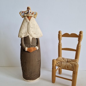 May include: A ceramic figurine of a woman wearing a crown and a white shawl, with a small wooden chair beside it. The figurine is brown with intricate cut-out designs. The chair has a woven seat.