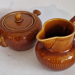 May include: A brown ceramic sugar bowl with a lid and a matching creamer jug. Both items have a ribbed texture and a glossy glaze. The sugar bowl features two handles and a knob on the lid.