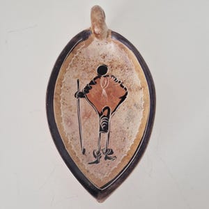 Peut inclure: Un plat en céramique en forme d'amande avec un bord brun foncé et un intérieur beige moucheté. Le centre présente une illustration graphique noire et terre cuite d'une silhouette tenant un bâton, entourée d'une bordure festonnée blanche.