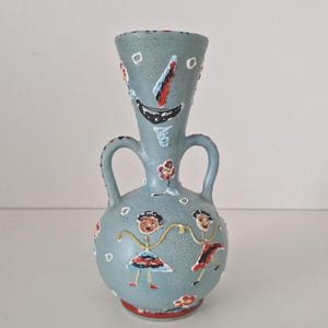 Pode incluir: Um vaso de cerâmica azul-claro com duas alças, apresentando uma superfície texturizada decorada com figuras pintadas à mão de duas pessoas de mãos dadas, pequenas flores vermelhas e brancas e formas abstratas em vermelho, branco e preto.