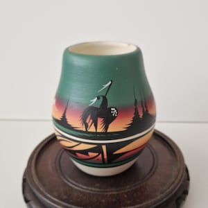 Op de afbeelding: Een kleine, ronde keramische vaas met een matte afwerking, beschilderd met een zwart silhouet van een paard tegen een kleurverloop van groen, oranje en rood. Het onderste gedeelte toont geometrische patronen in zwart en lichtbruin.