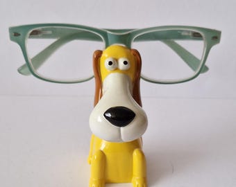 May include: A yellow dog-shaped spectacle holder with a white snout, brown ears, and black eyes. The holder supports a pair of light green spectacles. The spectacle holder is a decorative item.