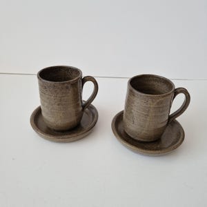 Pode incluir: Duas canecas de cerâmica a condizer com pegas, cada uma assente num pires circular. O conjunto apresenta um vidrado castanho escuro salpicado com estrias horizontais visíveis na superfície exterior das canecas, criando um aspeto terroso e texturado.