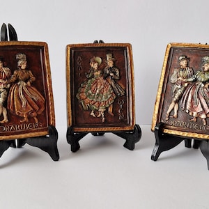 Könnte beinhalten: Drei rechteckige Reliefplatten, die Paare in traditioneller Kleidung zeigen, jeweils auf einem schwarzen Staffelei. Die Platten haben einen braunen Hintergrund mit goldfarbenen Rahmen und geprägten Figuren. Das Wort "VORARLBERG" ist auf jeder Platte zu sehen.