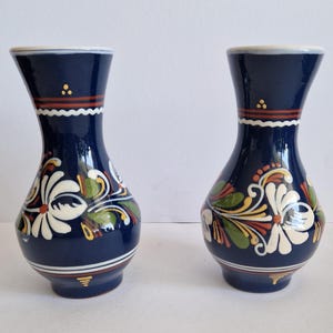 May include: Two navy blue ceramic vases with white floral designs, green leaves, and red and yellow accents. The vases have a flared top and a rounded base, with a white scalloped band near the top.