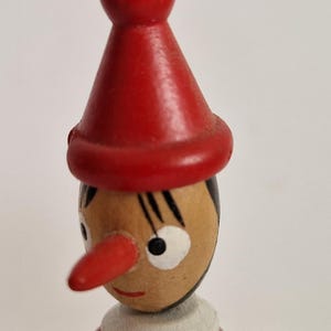 May include: A wooden Pinocchio figurine with a red hat and nose, black hair, and a white collar. The figure is painted in red, black, white, and tan. The figurine is a classic children's toy.