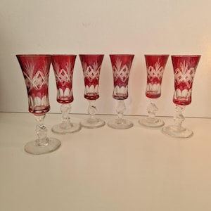 May include: A set of six tall cordial glasses featuring a deep red overlay cut to clear glass. Each glass has a flared bowl with a diamond and fan pattern, a clear twisted stem, and a circular base. The glasses are arranged in a semi-circle.