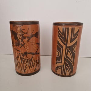 Könnte beinhalten: Zwei zylindrische Terrakotta-Vasen mit dunkelbraunen Rändern und Böden. Die linke Vase zeigt ein eingraviertes Weizenährenmuster vor dunklem Hintergrund. Die rechte Vase weist ein geometrisches Muster aus dunkelbraunen Linien auf Terrakotta auf.