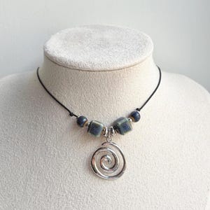 May include: A necklace featuring a silver-toned metal spiral pendant suspended from a black cord. The design includes two square-shaped blue and grey ceramic beads and two smaller round dark blue beads, accented with small silver-toned metal spacers.