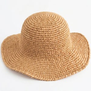 May include: A tan sun hat crafted from woven straw material. The hat features a rounded crown and a wide, flexible brim with a consistent, textured stitch pattern throughout the entire structure, creating a simple and functional head covering.