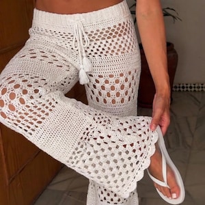 May include: White crochet pants featuring an openwork pattern, a drawstring waistband with two tassels, and flared legs with a scalloped hem. The pants are paired with white flip-flops, showcasing the intricate knit texture and design details.
