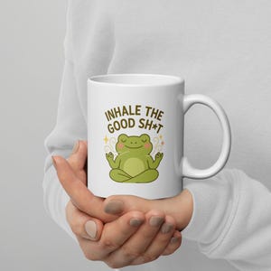 Peut inclure: Mug en céramique blanche avec un design humoristique. Le mug présente le texte "INHALE THE GOOD SH*T" au-dessus d'une grenouille de dessin animé en position de yoga. La grenouille est verte avec un visage souriant et est entourée d'étoiles décoratives.