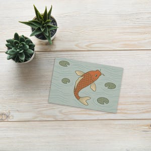 May include: A greeting card with an illustration of an orange koi carp in a light blue pond with lily pads. The card is on a light wood surface, with two small potted succulents. The design is minimalist.