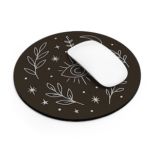 Puede incluir: Una alfombrilla de ratón redonda de color marrón oscuro con un ratón inalámbrico blanco. La alfombrilla presenta un diseño celestial con un ojo, estrellas y elementos botánicos en blanco. El ratón es blanco.
