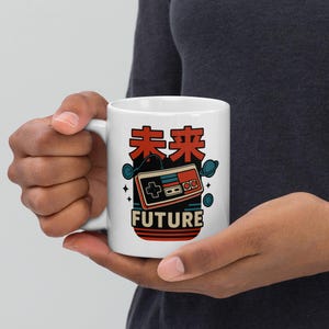 Può includere: Tazza in ceramica bianca con un design di controller di videogiochi retrò e la parola "FUTURE" in inglese, più caratteri giapponesi. Il design presenta elementi rossi, blu e neri, con un'estetica retrò.