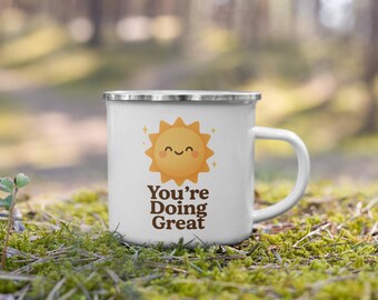 May include: White enamel mug with a silver rim and a white handle. The mug features a smiling sun graphic in orange and the text "You're Doing Great" in brown. The mug is set against a blurred green background.