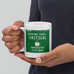 May include: White ceramic mug with a green graphic that reads "Caffeine Level: Critical" and "Administer Stim-Brew". The mug has a handle and is held by a person.