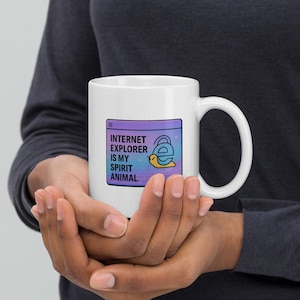 Könnte beinhalten: Weiße Keramik-Tasse mit dem Text "INTERNET EXPLORER IS MY SPIRIT ANIMAL" und einem Cartoon des Internet Explorer Logos. Das Design befindet sich auf einem lila und blauen Hintergrund. Die Tasse hat einen gebogenen Griff.