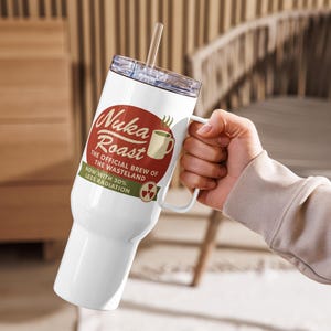 May include: White travel mug with a handle and a clear lid, featuring the text "Nuka Roast" in a retro design. The mug also includes the phrases "The Official Brew of the Wasteland" and "Now with 30% Less Radiation."
