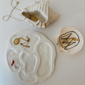 Op de afbeelding: Een verzameling witte decoratieve trays en schelpvormige containers. De trays bevatten gouden kettingen en een rode chilipeper hanger ketting. Eén schelpvormige container heeft een gouden ketting met een hanger. De andere heeft een zwart-goud ontwerp.