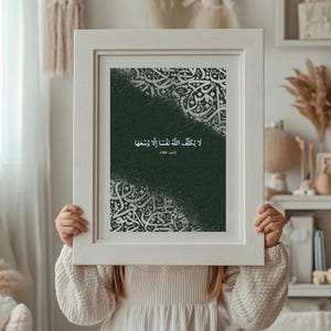 Puede incluir: Una impresión enmarcada con caligrafía árabe blanca sobre un fondo verde oscuro. La persona sostiene la impresión. El marco es blanco. El texto de la impresión está en árabe.