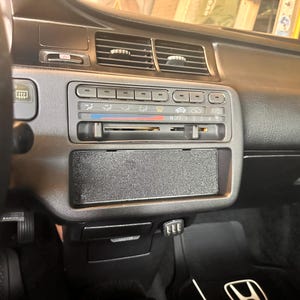 May include: A black car dashboard featuring air vents, climate controls, and a radio slot. The dashboard has a textured surface and a small storage compartment. The steering wheel is visible on the left side of the frame.