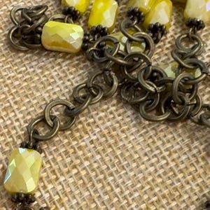 May include: A gold chain necklace with yellow faceted glass beads. The necklace is on a brown woven surface.