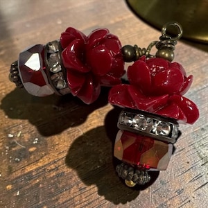May include: Two decorative charms featuring deep red carved resin roses, clear faceted crystal beads, and metallic spacers. Each piece is finished with a small bronze-toned metal loop at the top for attachment, showcasing a detailed floral design.