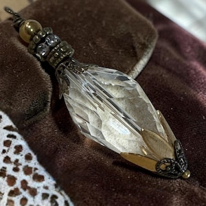 May include: A clear, faceted crystal pendant with ornate metalwork. The pendant has a teardrop shape with gold-toned accents and a decorative top with small, sparkling stones. The piece is likely a decorative element or jewelry.