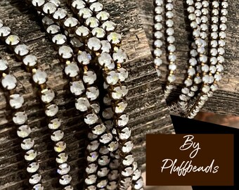 Puede incluir: Primer plano de una cadena multi-hilo hecha de pequeños diamantes de imitación transparentes. La cadena está sobre una superficie de madera marrón. El texto "By Plufffbeads" es visible en la esquina inferior derecha de la imagen.