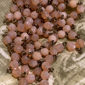 以下が含まれることがあります： 真鍮のチェーンに、薄いピンク色のファセットカットのガラスビーズが連なっています。ビーズは小さく、わずかに虹色に光る仕上げになっています。