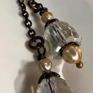 May include: A pair of vintage-style chandelier earrings. The earrings feature faceted clear crystal pendants, cream-colored pearl-like beads, and antique bronze-colored chain links. The earrings are designed to dangle.