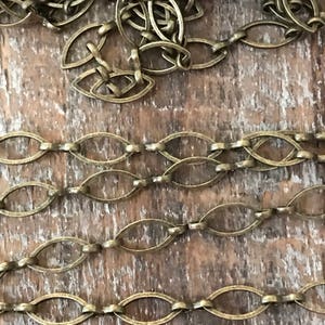 May include: A close-up of a chain made of oval-shaped links. The chain is made of a brass-colored metal and is lying on a wooden surface.