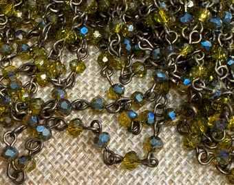 May include: A close-up of a chain of dark brown metal links with olive green faceted glass beads. The chain is lying on a woven fabric surface.