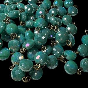 May include: Close-up of a teal-colored beaded necklace. The beads are faceted, giving them a sparkling appearance. The necklace is strung together with gold-colored metal links, creating a vintage or antique aesthetic. The background is black.