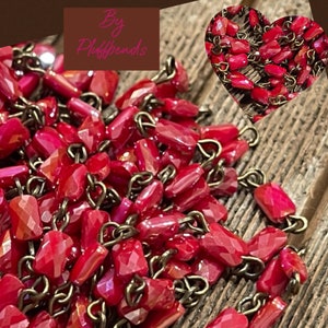 May include: A close-up of a red faceted bead chain necklace with antique brass chain links. The beads are strung on the chain and are connected to each other with small loops. The chain is lying on a wooden surface. The text "By Pluffbeads" is visible in the top left corner of the image.