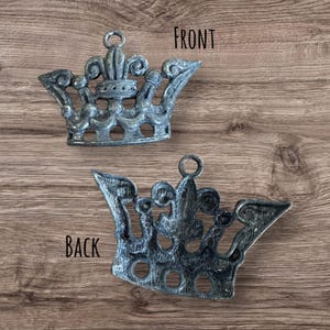 May include: A silver metal crown ornament with a fleur-de-lis design. The front and back of the ornament are shown in the image. The ornament is on a wooden background.