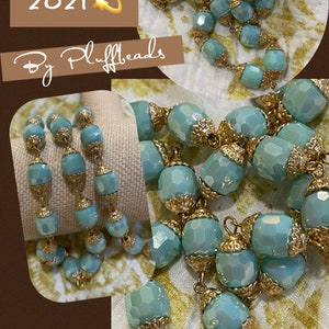 May include: A close-up of a turquoise blue glass bead necklace with gold accents. The beads are faceted and have a slight iridescent sheen. The necklace is new for 2021 and is by Pluffbeads.