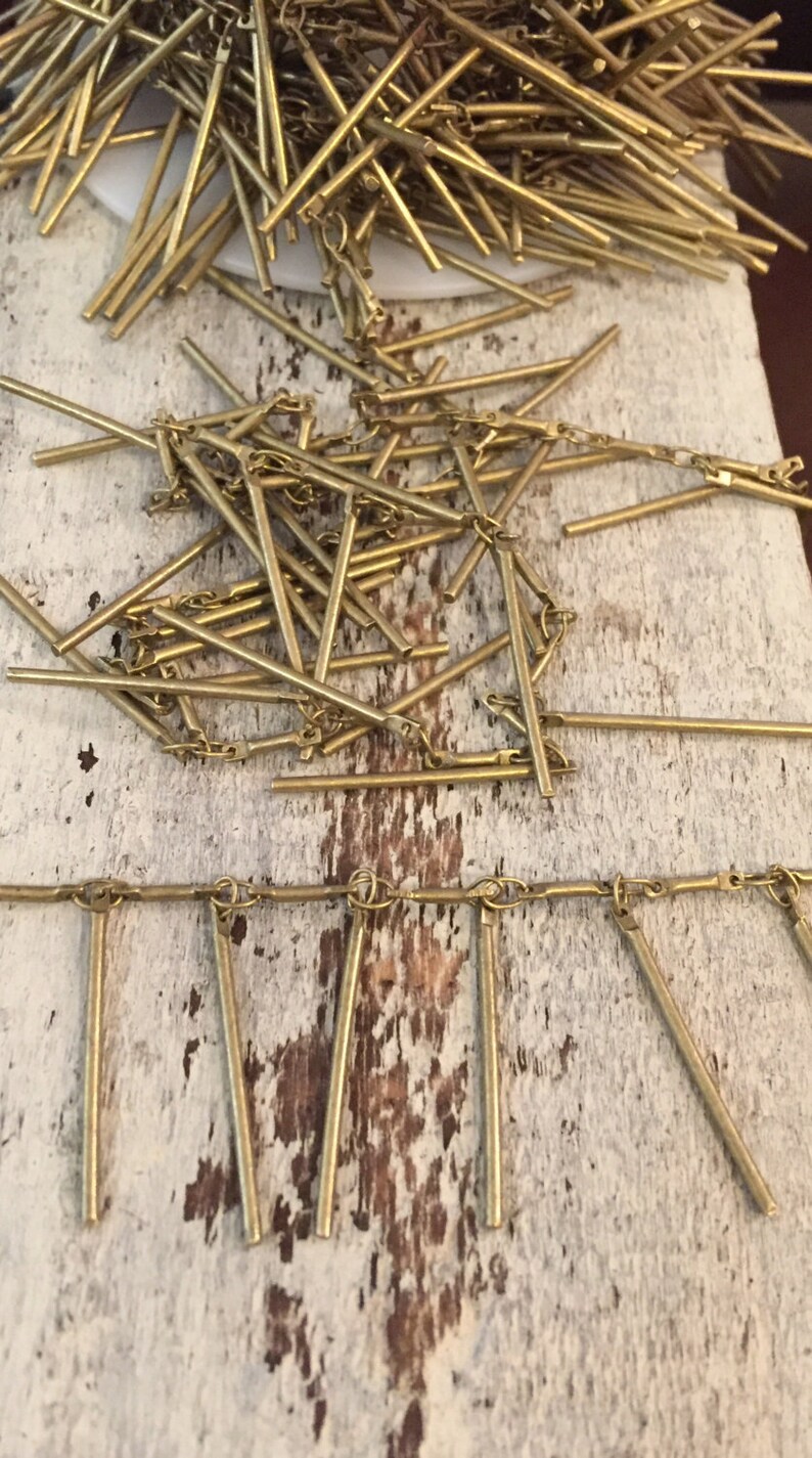 May include: A gold chain with multiple gold metal rods hanging from it. The chain and rods are on a white wooden surface.