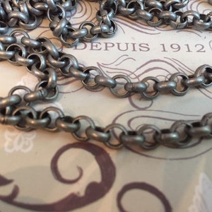 Pode incluir: Um colar de corrente de prata com um design simples e ligado. A corrente est&aacute; sobre uma superf&iacute;cie estampada com o texto "DEPUIS 1912".