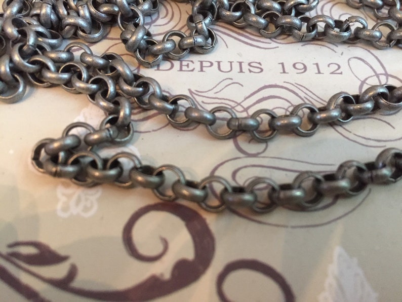 Pode incluir: Um colar de corrente de prata com um design simples e ligado. A corrente est&aacute; sobre uma superf&iacute;cie estampada com o texto "DEPUIS 1912".