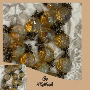 Könnte beinhalten: Nahaufnahme einer Halskette mit facettierten, irisierenden, bernsteinfarbenen Perlen. Die Kugeln sind kugelförmig und durch antike, messingfarbene Metallglieder verbunden. Der Text "By Pluffbeads" ist unten rechts sichtbar.
