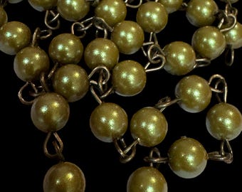 Op de afbeelding: Close-up van een ketting met olijfgroene, ronde kralen. De kralen zijn met kleine, metalen schakels aan elkaar geregen. De ketting is geplaatst tegen een zwarte achtergrond, waardoor de kleur en textuur van de kralen worden benadrukt.