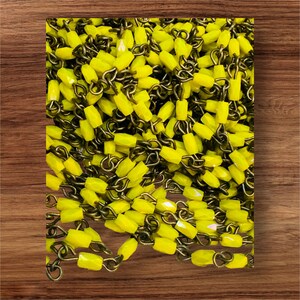 May include: A close-up shot of a pile of yellow, faceted beads strung on antique brass-colored chains. The beads are oblong and have a slight sparkle. The chains are looped together, creating a dense, textured pattern. The background is a wooden surface.