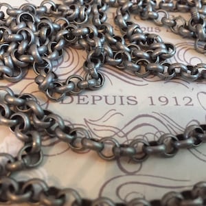 Pode incluir: Uma corrente de prata com um design de elo grosso e arredondado. A corrente est&aacute; sobre um peda&ccedil;o de papel com o texto "DEPUIS 1912" impresso nele.