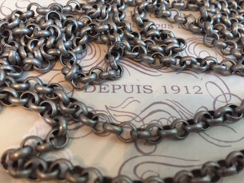 Pode incluir: Uma corrente de prata com um design de elo grosso e arredondado. A corrente est&aacute; sobre um peda&ccedil;o de papel com o texto "DEPUIS 1912" impresso nele.