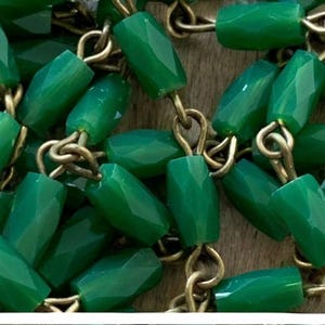May include: A close-up of a necklace with faceted, emerald-green beads. The beads are strung together with gold-colored metal links. The necklace is displayed on a light brown wooden surface.