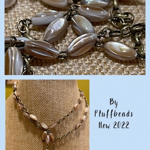 May include: A double-strand necklace with a brown chain and oval-shaped iridescent shell beads. The necklace has a long pendant with a tassel of shell beads. The necklace is on a brown fabric background. By Pluffbeads New 2022.