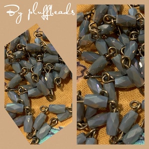 May include: Close-up of a collection of faceted, light blue beads strung together with antique-style metal loops. The beads are arranged in a diagonal pattern against a textured, tan background. The text "By pluffbeads" is in the upper left corner.