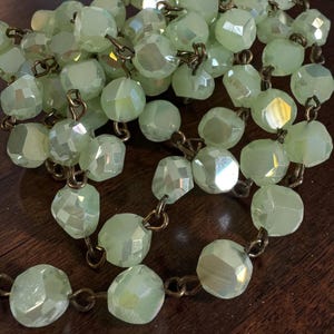 May include: A necklace featuring light green, faceted beads strung on a bronze-colored chain. The beads have an iridescent quality, reflecting multiple colors. The necklace is displayed on a dark brown surface.
